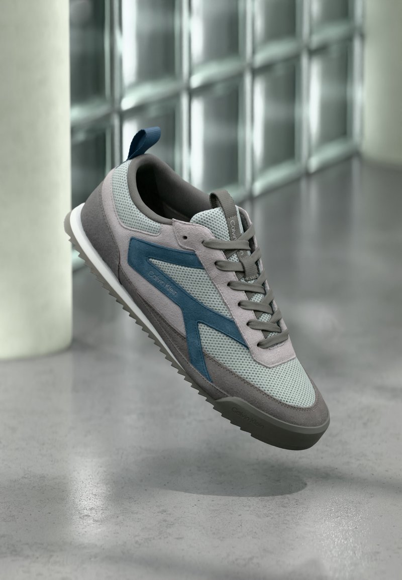 Γκρι και μπλε sneaker Calvin Klein με λεπτομέρειες από δίχτυ και σουέντ, αιωρούμενο πάνω από ένα γυαλισμένο τσιμεντένιο πάτωμα κοντά σε έναν τοίχο από γυάλινους κύβους.