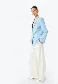 Lichtblauwe blazer met klepels, dubbele knopen en zijzakken, gecombineerd met off-white wijde broek en puntige schoenen.