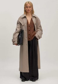 Beige wollen overcoat met grote kraag, knopen aan de voorkant, gedragen over een bruine, harige cardigan en zwart wijde broek. Houdt een zwarte handtas vast.