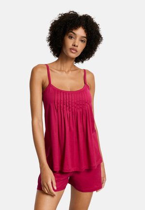 Kobieta o kręconych włosach, ubrana w bezrękawnik plisowany top w kolorze fuksji oraz dopasowane szorty, stojąca na jednolitym tle.