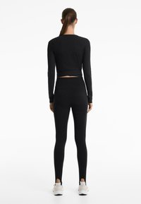 Woman standing facing away, wearing black long-sleeve top, high-waisted black leggings with heel cutouts, and white sneakers on a white background.