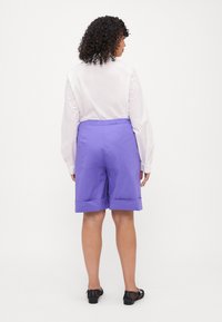 Pantaloni corti viola con cintura elastica, con orlo risvoltato, abbinati a una camicia bianca con bottoni, visti da dietro.