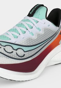 Λευκό και ανοιχτό τιρκουάζ παπούτσι τρεξίματος με μπορντό και πορτοκαλί λεπτομέρειες, λευκά κορδόνια και επάνω μέρος από δίχτυ που δείχνει το λογότυπο της μάρκας στην πλευρά.