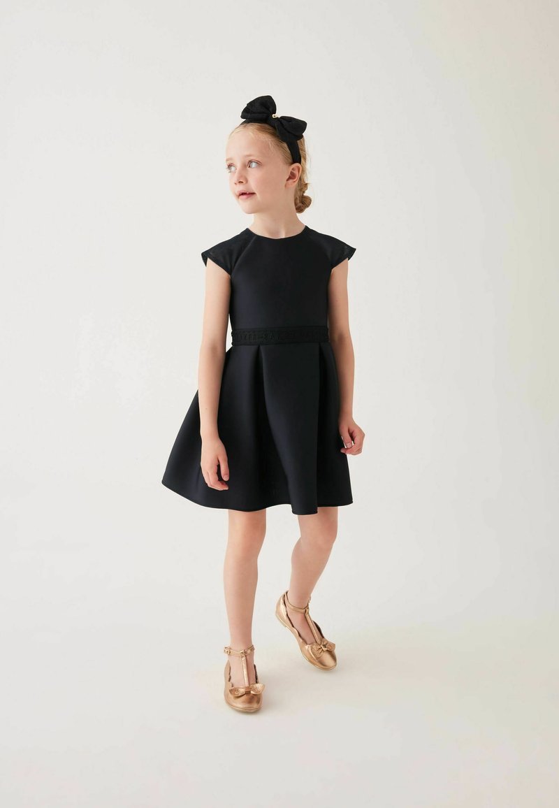 Schwarzes ärmelloses Kleid mit einem ausgestellten Rock, strukturiertem Taillenbund, kombiniert mit goldenen T-Riemchen-Schuhen und einem schwarzen Schleifen-Stirnband.