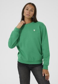 Grüner Pullover aus weichem Material mit gerippten Bündchen und Saum, auf der linken Brust mit einem kleinen weißen Herz-Logo versehen. Getragen mit schwarzen Hosen.