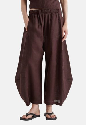 Brune vidde cropped bukser med elastisk talje og sidelommer, båret med sorte klip-klappere på bare fødder, vist fra taljen til tæerne.