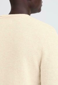 Light beige sweater with a textured, knitted fabric. Features a ribbed neckline and seams, showcasing a simple design without patterns.