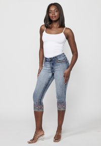 Blaue Denim-Caprijeans mit dekorativen Nähten am Saum, mit einer taillierten Passform und fünf Taschen. Kombiniert mit einem weißen Tanktop.