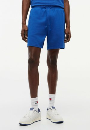 Man draagt blauwe short met trekkoord, witte crew sokken met logo en witte sneakers tegen een effen achtergrond.