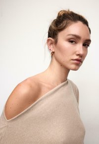 Bež pulover brez ramen z mehko teksturo. Ima sproščen kroj in diskretne šive. Model nosi zlate uhančke obročke z okrasnimi kamni.