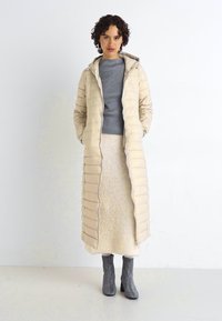 Beige, lang polstret frakk med hette og vertikal quiltedesign. Bær over en grå topp og kremfarget skjørt, kombinert med grå hæler.
