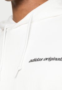 Vit hoodie i mjukt material med dragsko och en svart "adidas originals"-logotyp tryckt på framsidan.