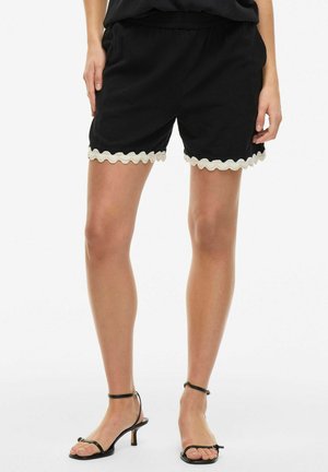 Black shorts with an elastic waistband, featuring a scalloped cream trim at the hem. Smooth fabric and relaxed fit. Ankle strap sandals visible.