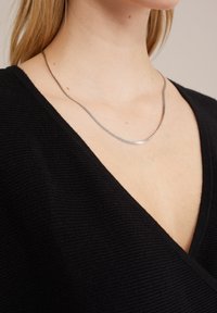 Kovový náhrdelník s elegantním řetízkovým designem. Nošený přes černou strukturovanou tkaninu s výstřihem do V. Barevný kontrast mezi stříbrnou a černou.