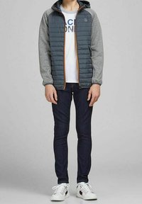 Szara kurtka puchowa z kontrastowymi rękawami i kieszeniami bocznymi, noszona na białym podkoszulku z grafiką, w połączeniu z ciemnymi dżinsami i białymi sneakersami.