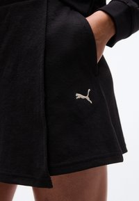 Κοντινό πλάνο ενός ατόμου που φοράει μια μαύρη υφασμάτινη φούστα με λευκό λογότυπο Puma και το χέρι του ακουμπισμένο στην πλαϊνή τσέπη.