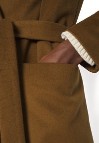 Cappotto di lana marrone con cintura annodata, caratterizzato da una tasca laterale e dettagli in polsino a costine color avorio, che mostrano un tessuto morbido e strutturato.