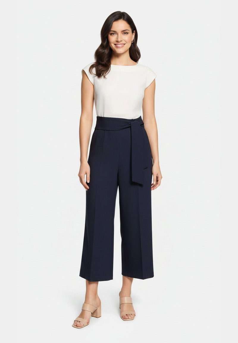Femme aux longs cheveux foncés portant un haut blanc sans manches, un pantalon large court bleu marine avec une ceinture, et des sandales beiges à talons carrés, debout en souriant.