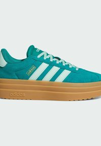 Baskets Adidas en daim sarcelle avec bandes blanches, lacets bleu clair et semelle épaisse en caoutchouc beige sur fond uni clair.