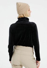 Top lungo in velluto nero con colletto alto, abbinato a pantaloni beige a vita alta con dettagli cuciti e logo discreto sul retro.