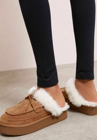 Zapatillas de ante en color marrón claro con ribete de piel sintética blanca, que cuentan con una suela plana y detalles decorativos de lazo y costuras.