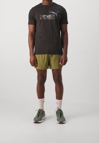 Černé tričko s krátkým rukávem s šedým logem Puma, doplněné olivově zelenými šortkami, bílými ponožkami a zelenými běžeckými botami s hnědými akcenty.