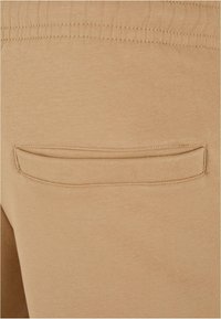 Beige Jogginghose aus weichem Material, mit einem elastischen Bund und einer einzelnen Gesäßtasche mit gebogenem Klappdeckel.