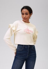 Felpa crema cropped con maniche a volant e grafiche rosse della Torre Eiffel con la scritta "Parigi". Indossata con jeans blu a vita alta.