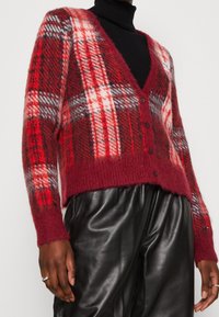 Cardigan à carreaux rouges et noirs avec un col en V, fermeture à boutons et texture duveteuse, associé à un pantalon en cuir noir.