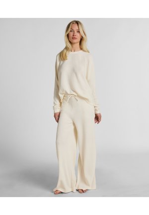 Vrouw staat op blote voeten, draagt een losse, crèmekleurige, wafelgebreide lange mouw top en bijpassende wijde broek, tegen een effen grijze achtergrond.