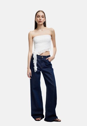 Slanke vrouw in een witte strapless ruches crop top, donkere wijde jeans en zwarte sandalen, staand met één hand in de zak.