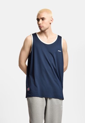 Młody mężczyzna z krótkimi blond włosami, ubrany w granatową sportową koszulkę bez rękawów i jasnoszare spodnie dresowe na jednolitym tle.