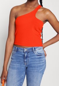 Červený žebrovaný crop top s jedním ramínkem, který má přiléhavý střih. Spárován se světle modrými džíny a oranžovým doplňkem.