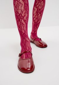 Zapatos planos de cuero rojo con una correa y hebilla, combinados con unas medias de encaje fucsia con un patrón floral.