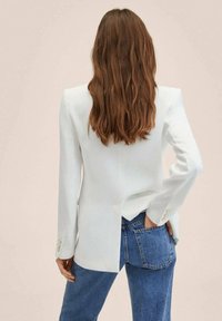 Kobieta z długimi brązowymi włosami, ubrana w białą marynarkę i niebieskie dżinsy, stoi z jedną ręką w tylnej kieszeni, zwrócona plecami do aparatu.