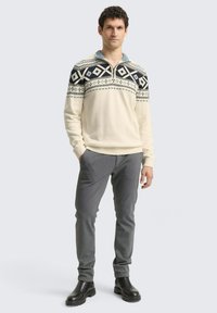 Cremfarbener Strickpullover mit schwarzem geometrischem Muster an der Passe, Reißverschlusskragen und grauen Hosen. Das Modell trägt schwarze Stiefeletten.