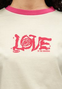 Ανοιχτό μπεζ μπλουζάκι με ροζ ribbed γιακά που έχει έντονο κόκκινο κείμενο "LOVE" και ένα σπειροειδές γραφικό, μαζί με το κείμενο "OF THE UNKNOWN" από κάτω.