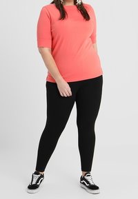 Kvinna i korallfärgad kortärmad topp, svarta leggings och svartvit sneakers som står mot en enkel ljus bakgrund.