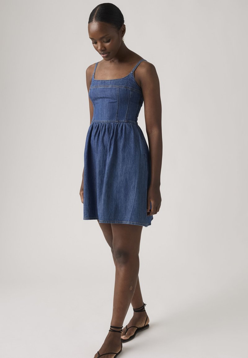 Woman in a sleeveless blue denim dress and black sandals looking down, standing against a plain light background.