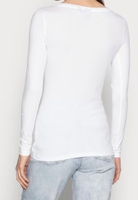 Kvinna som bär en åtsittande vit långärmad skjorta och ljusblå denimjeans, visad bakifrån mot en plain bakgrund.