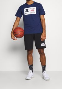 Muž v námořnickém tričku Champion a černých šortkách držící basketbal Nike, s černými ponožkami Nike a bílými teniskami, stojící na šedé podlaze.