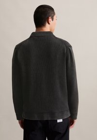 Donkergrijze gebreide jack met een ribbelkraag en textuurstof. Zichtbare stiknaaddetail op de schouders en een label aan de zoom.