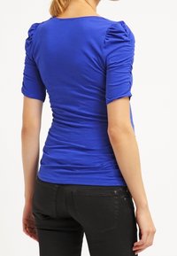 Top ajustado de color azul royal con mangas cortas abullonadas; hecho de una tela elástica, con detalle de fruncidos a lo largo de los lados y la parte posterior.