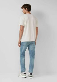 Young man wearing a plain white short-sleeve shirt, light blue jeans, and white sneakers standing facing a gray wall.
