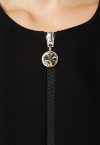 Tissu noir avec une fermeture éclair frontale dotée d'un curseur argenté et d'un accent de gemme ronde transparente en bas. Texture lisse, design élégant.