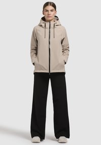 Beige lätt jacka med huva, frontdragkedja och svarta accenter, kombinerad med vida svarta byxor och vita sneakers. Enkel, modern design.