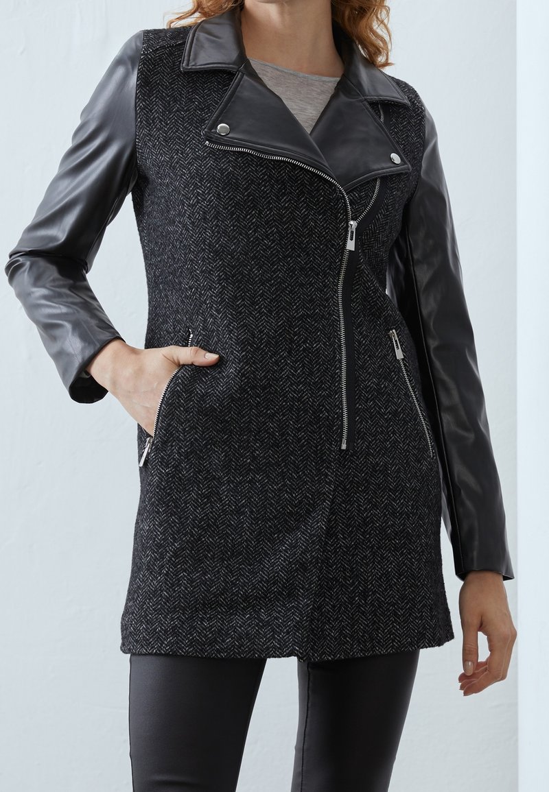 Kabát ve stylu motorkáře se všitým tělem z černé tkaniny vzoru rybí kost a lesklými černými koženými rukávy. Má zipové zapínání a dvě boční kapsy.