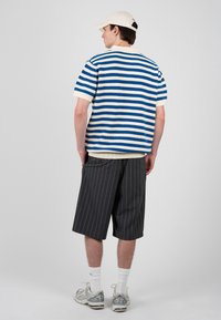 Hombre con gorra blanca, suéter de manga corta a rayas azules y blancas, pantalones cortos oscuros con rayas finas hasta la rodilla, calcetines blancos y zapatillas plateadas, de espaldas.