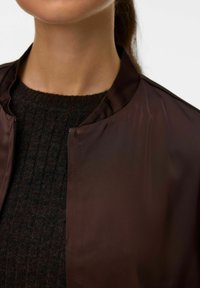 Giubbotto bomber marrone con una texture lucida e un colletto morbido, indossato sopra un maglione a coste scuro. I dettagli della zip sono visibili sul giubbotto.