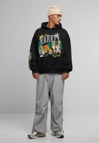 Zwarte hoodie met kleurrijke campingafbeelding, oversized pasvorm en lange mouwen. Lichtgrijze, losse broek en zilveren sportschoenen.